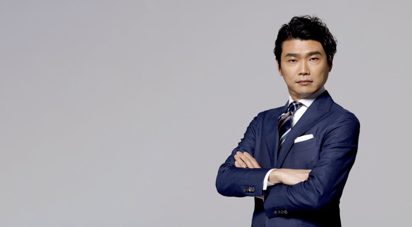 A Japanese man in a sharp business suit standing in front of a grey background, with his arms folded looking directly at the camera, his body tilted away at 3/4. 