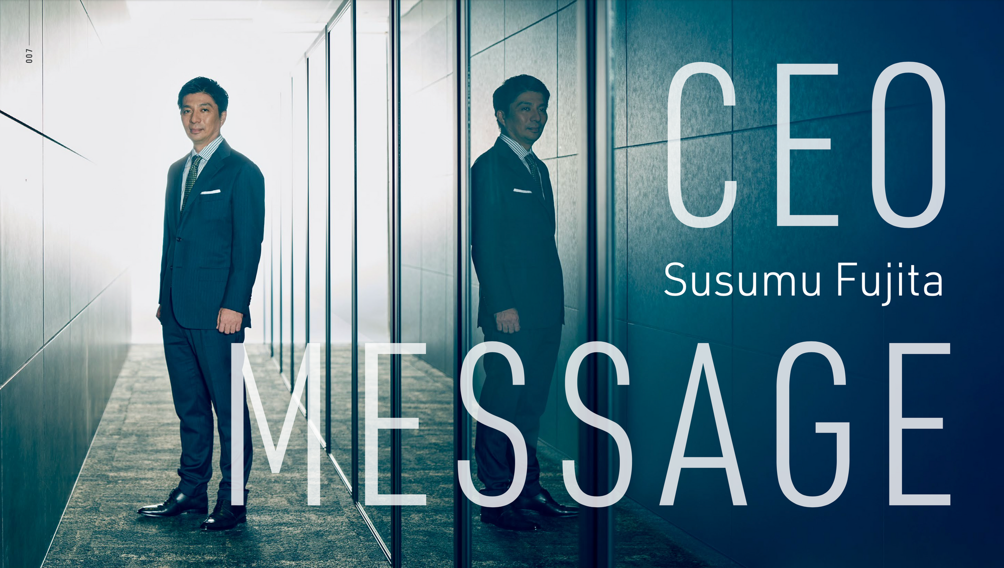 A Japanese man in a suit stars at the camera in a serious pose in a corridor, with his reflection on the right