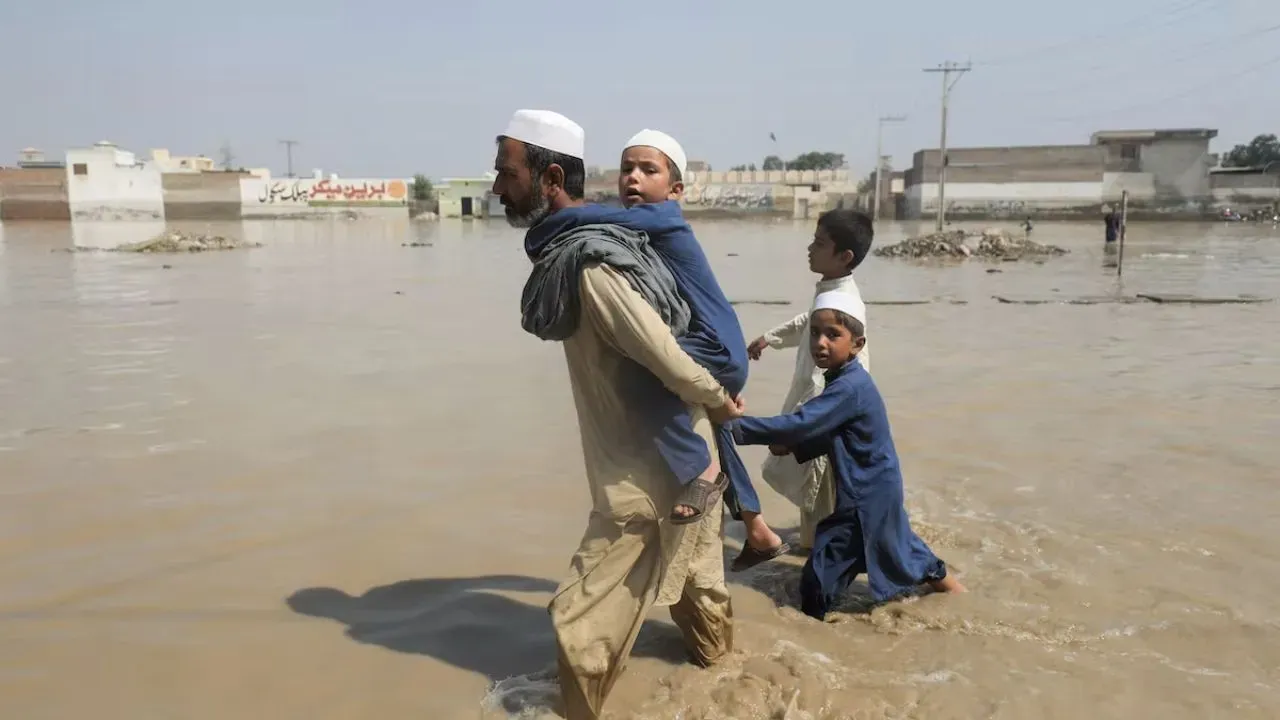 A man walks through muddy flood waters water with a child on his back, leading two other children.
