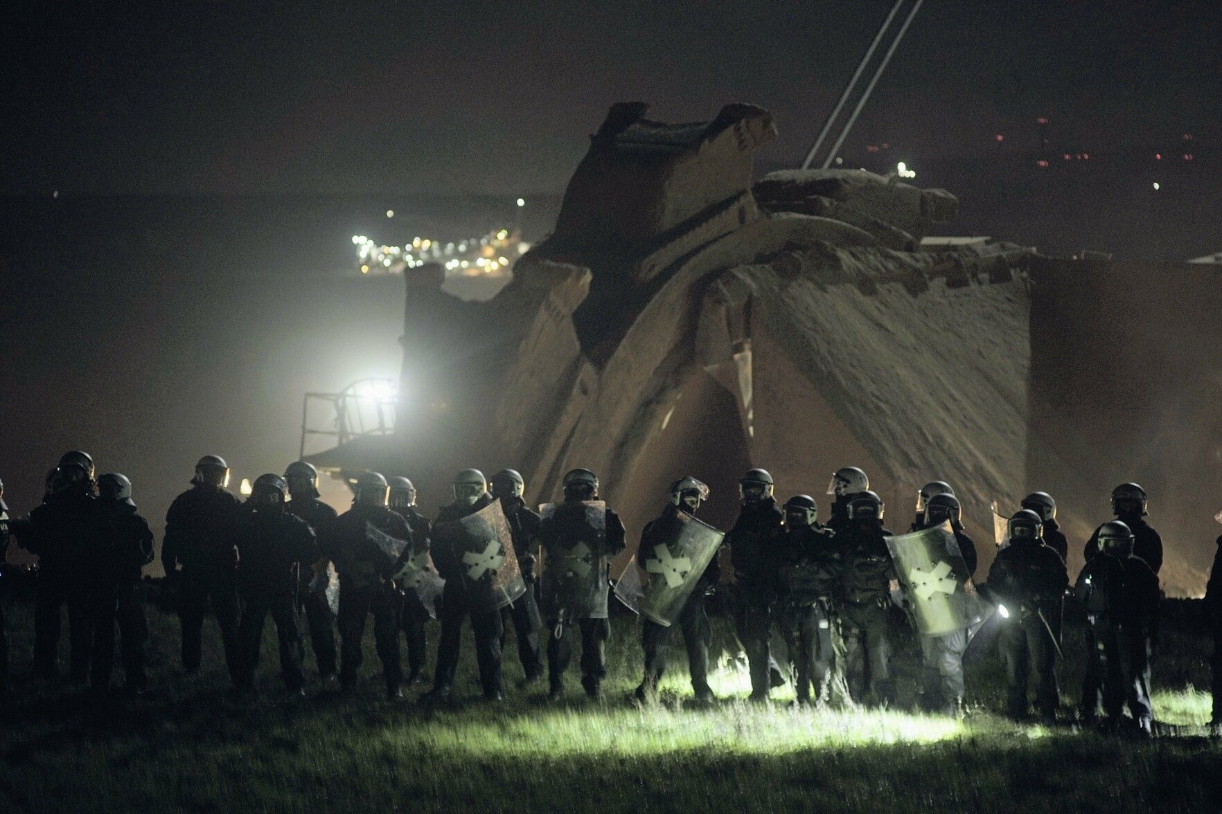 German riot police in full armour stand in front of a coal mining machine at night, lit by floodlights