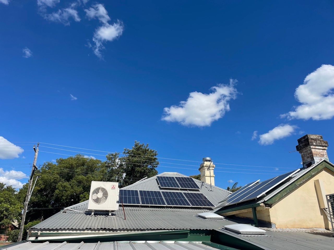 Several rows of solar panels on the roof of a house, trees and a telegraph pole behind it, a large bright blue sky above.