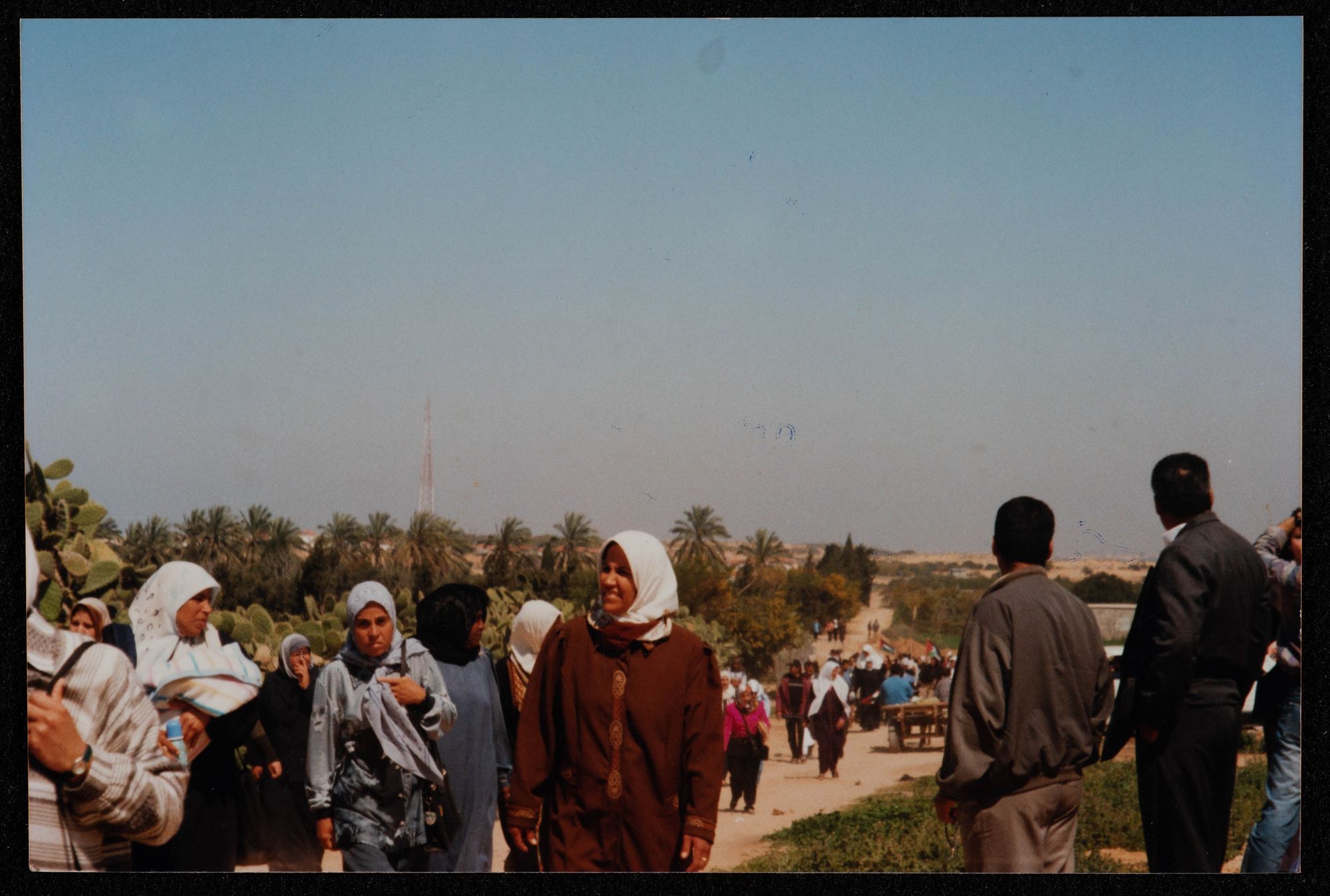 Women in headscarves approach up a dusty road, men with their backs to the camera look down it to palms in the distance