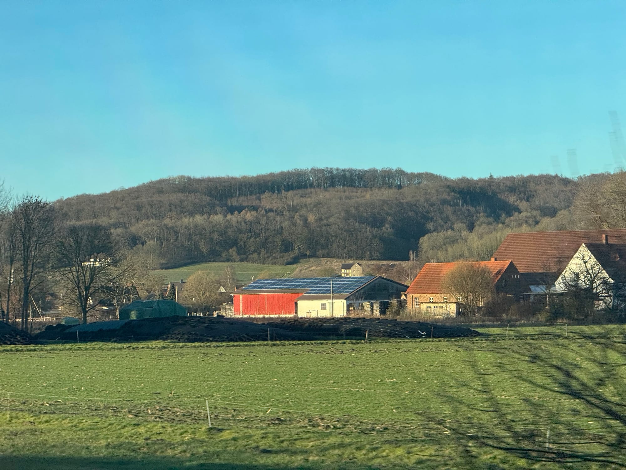 A German house covered in solar panels, a green field in the foreground, and a tree covered hill behind