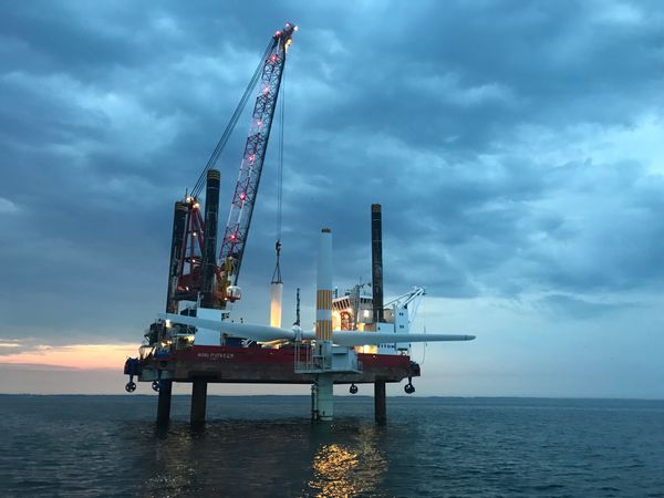 A raised platform sits above the ocean at dusk. A crane has lifted parts of a wind turbine off its pylon.