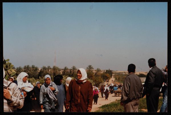 Women in headscarves approach up a dusty road, men with their backs to the camera look down it to palms in the distance