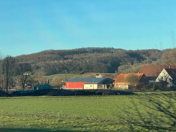 A German house covered in solar panels, a green field in the foreground, and a tree covered hill behind
